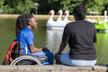 Teenage girl in wheelchair with friend relaxing by pond in park