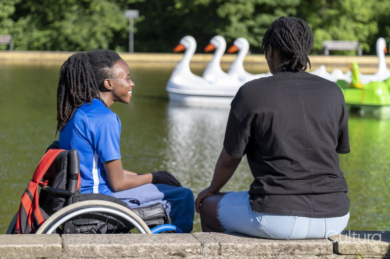 Teenage girl in wheelchair with friend relaxing by pond in park