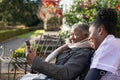 Smiling nurse and senior woman in wheelchair relaxing in park and looking at phone