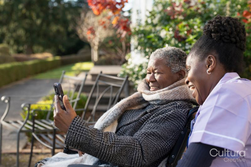 Smiling nurse and senior woman in wheelchair relaxing in park and looking at phone