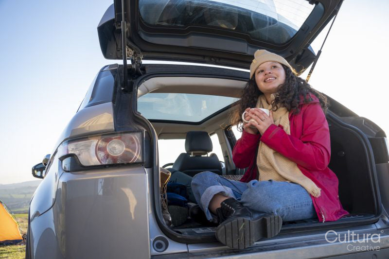 Young woman sitting in car trunk and drinking tea