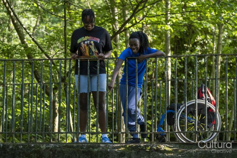 Teenage girl in wheelchair with friend standing on bridge in park