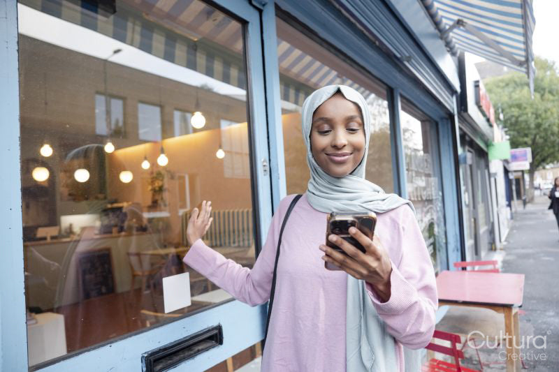 Young woman in hijab using phone in front of cafe