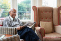Smiling senior man sitting in armchair and reading newspaper in nursing home