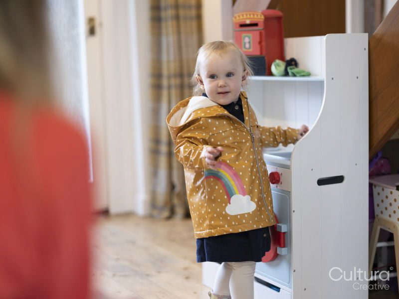 Blond girl wearing hearing aids and raincoat standing by toy kitchen at home