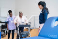 Female therapist and nurse assisting senior woman walking with mobility walker in exercise room