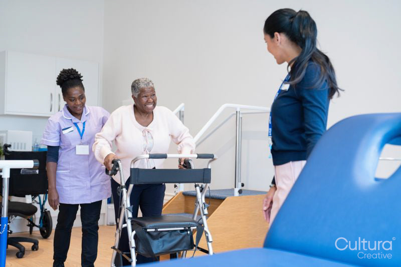 Female therapist and nurse assisting senior woman walking with mobility walker in exercise room