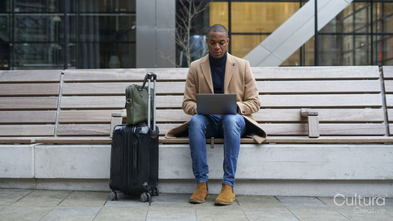 Business man with suitcase sitting on bench using laptop