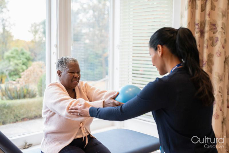 Female therapist assisting senior woman exercising with ball