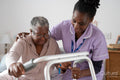 Smiling caregiver assisting senior woman sitting on bed and holding mobility walker