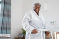 Smiling senior woman in bathrobe standing with mobility walker in bedroom