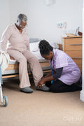 Caregiver helping senior woman sitting on bed to put on slippers