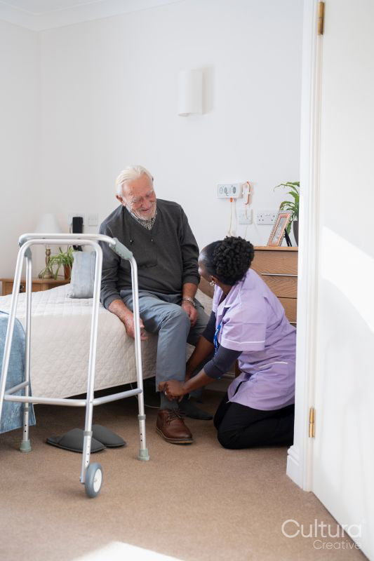 Caregiver helping senior man sitting on bed to put on shoes