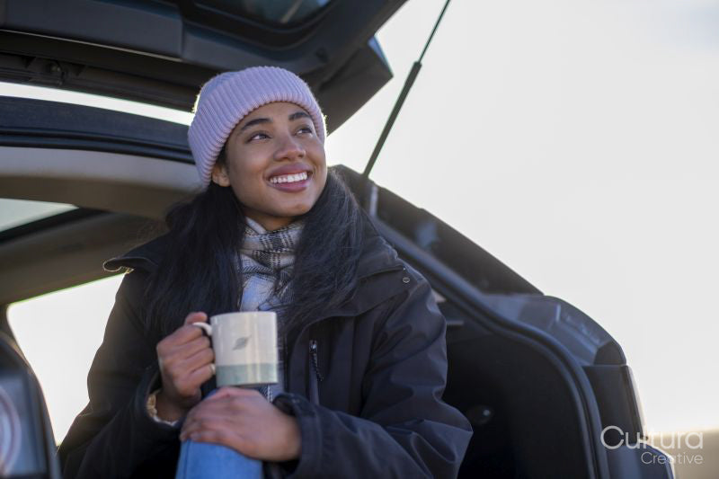 Young woman sitting in car trunk and holding mug
