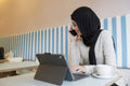 Young woman in hijab working on tablet and talking on phone in cafe
