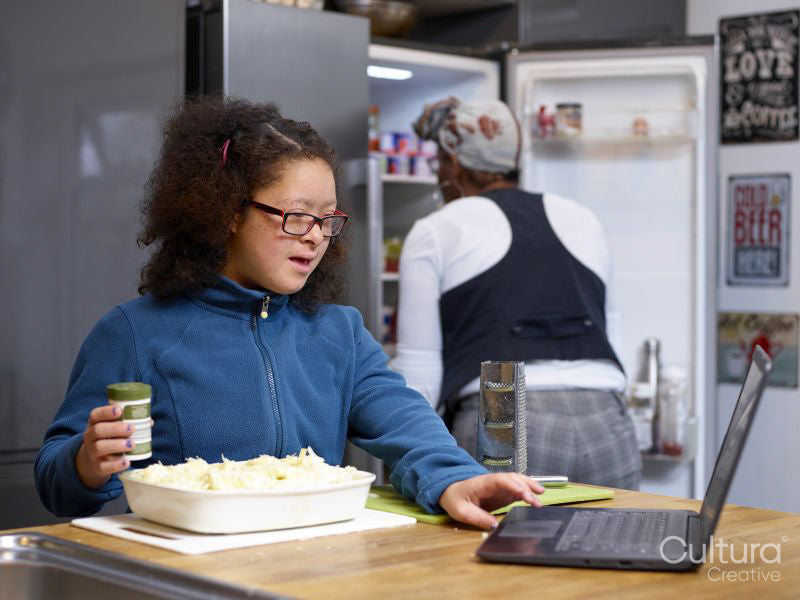 Girl with Down Syndrome using laptop and preparing food in kitchen