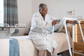Senior woman in bathrobe sitting on bed and holding mobility walker in nursing home