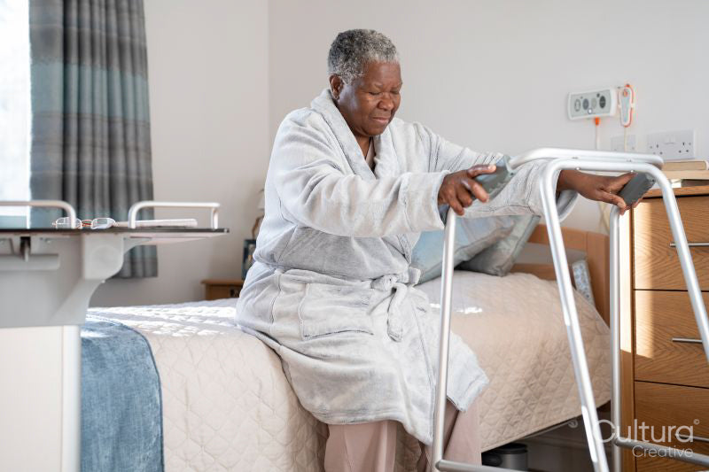Senior woman in bathrobe sitting on bed and holding mobility walker in nursing home