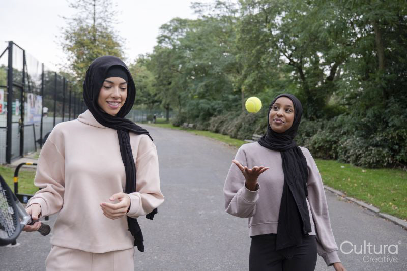 Two women in hijabs walking in park with tennis racket and ball