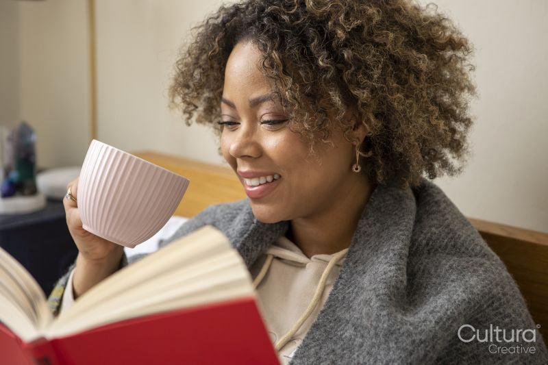 Smiling woman wrapped in shawl reading book and holding cup