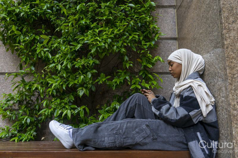 Young woman wearing hijab sitting on bench, looking at smart phone