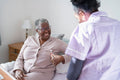 Caregiver assisting smiling senior woman sitting on bed, holding remote control
