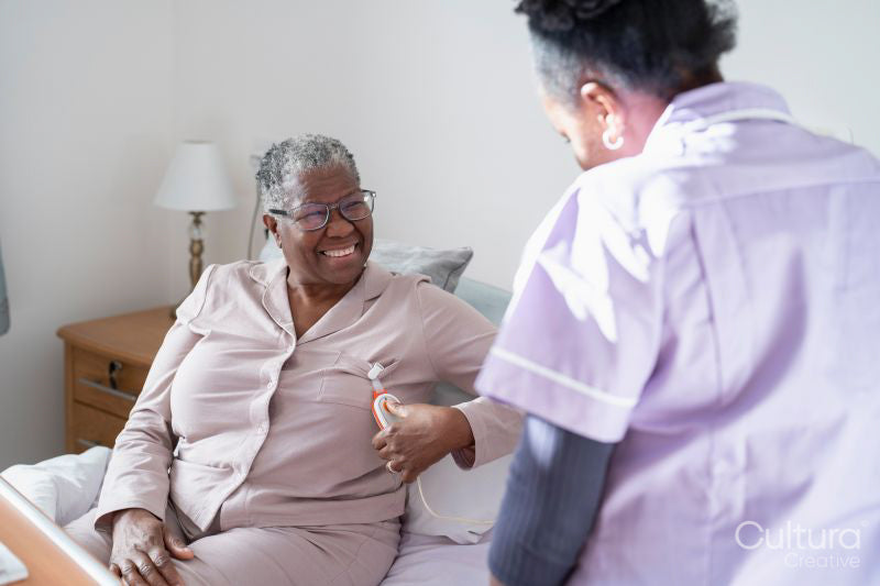 Caregiver assisting smiling senior woman sitting on bed, holding remote control
