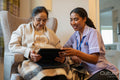 Senior lady sitting in armchair and showing tablet to nurse at nursing home