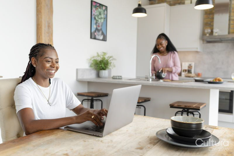 Woman working on laptop while girlfriend preparing breakfast in kitchen