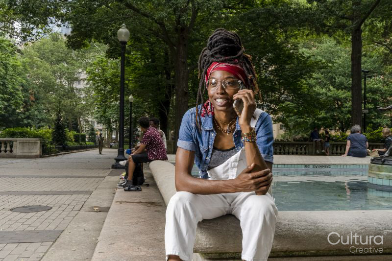 Portrait of young woman sitting on wall in park, talking on smart phone