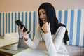 Smiling young woman in hijab using phone in cafe