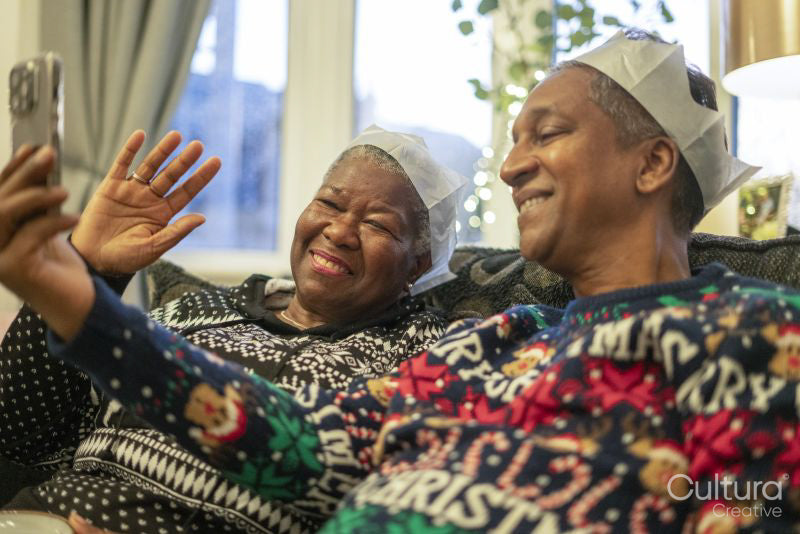 Senior couple sitting on sofa wearing Christmas decorations and waving to phone
