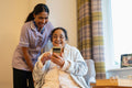 Senior lady looking at phone with nurse in room at nursing home