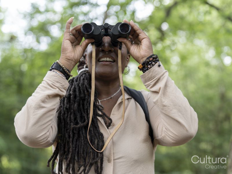 Smiling female hiker looking through binoculars in forest