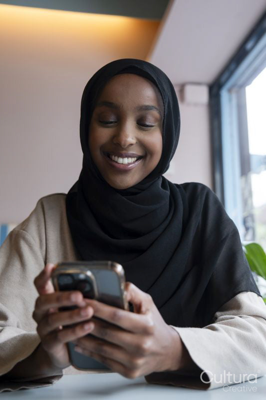 Smiling young woman in hijab using phone in cafe