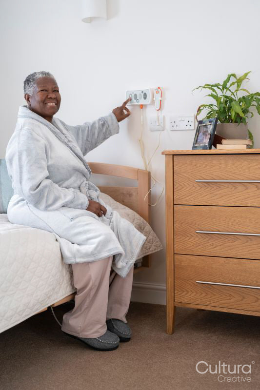 Smiling senior woman in bathrobe sitting on bed and pushing emergency button on wall