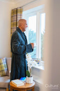 Senior man standing with coffee and looking through window in his room at nursing home