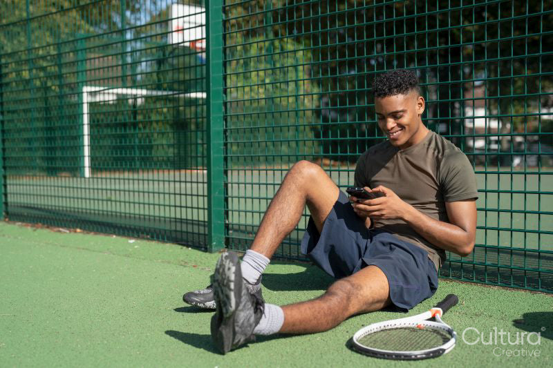 Smiling man sitting in tennis court, looking at smart phone