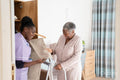 Smiling caregiver assisting senior woman standing at mobility walker and choosing pants at closet