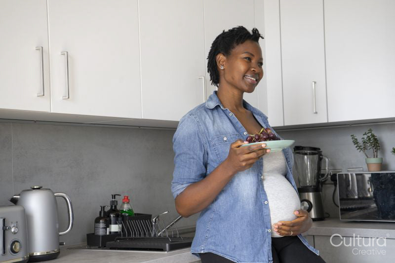 Smiling pregnant woman holding plate with red grapes in kitchen