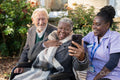 Smiling senior people accompanied by nurse relaxing in park and looking at phone