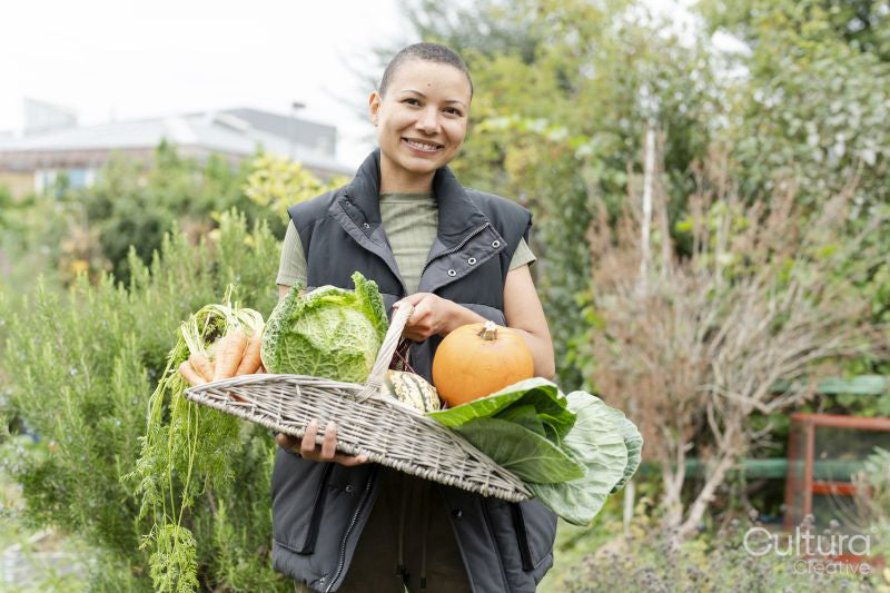 Portrait of smiling woman holding basket with fresh vegetables in urban garden